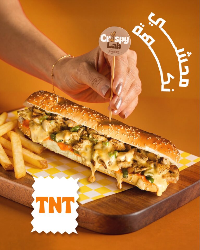 A hand pushing a Crispy Lab flag pick into the TNT chicken sub, served on a wooden board with fries.